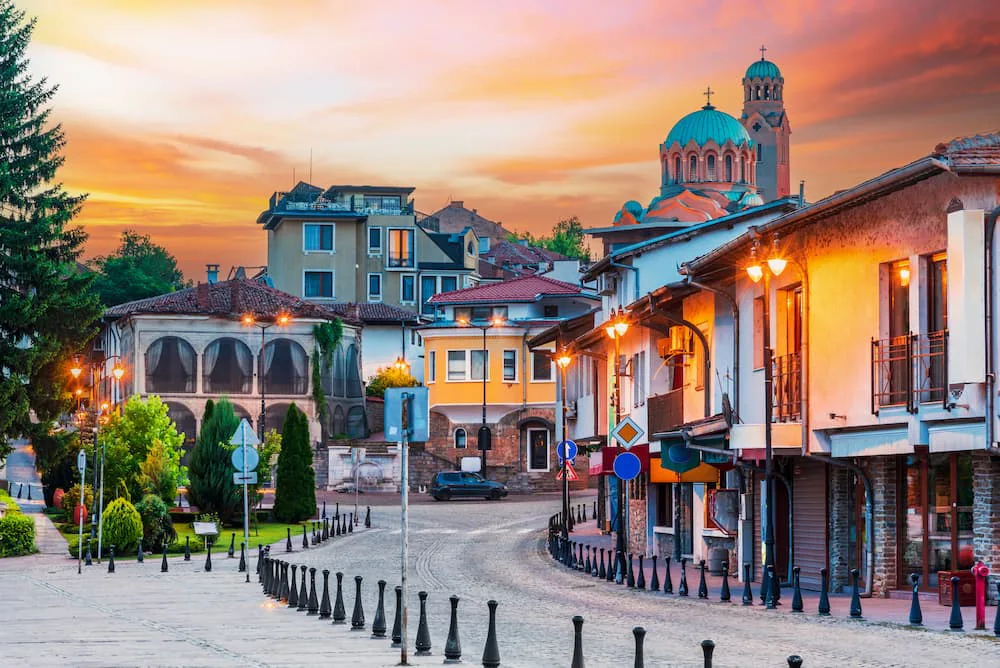 A quiet cobblestone street in the Old Town of Plovdiv, Bulgaria, lined with traditional pastel-colored houses and warm streetlights at sunset, with historic architecture and a domed church rising in the background under an orange and pink sky.