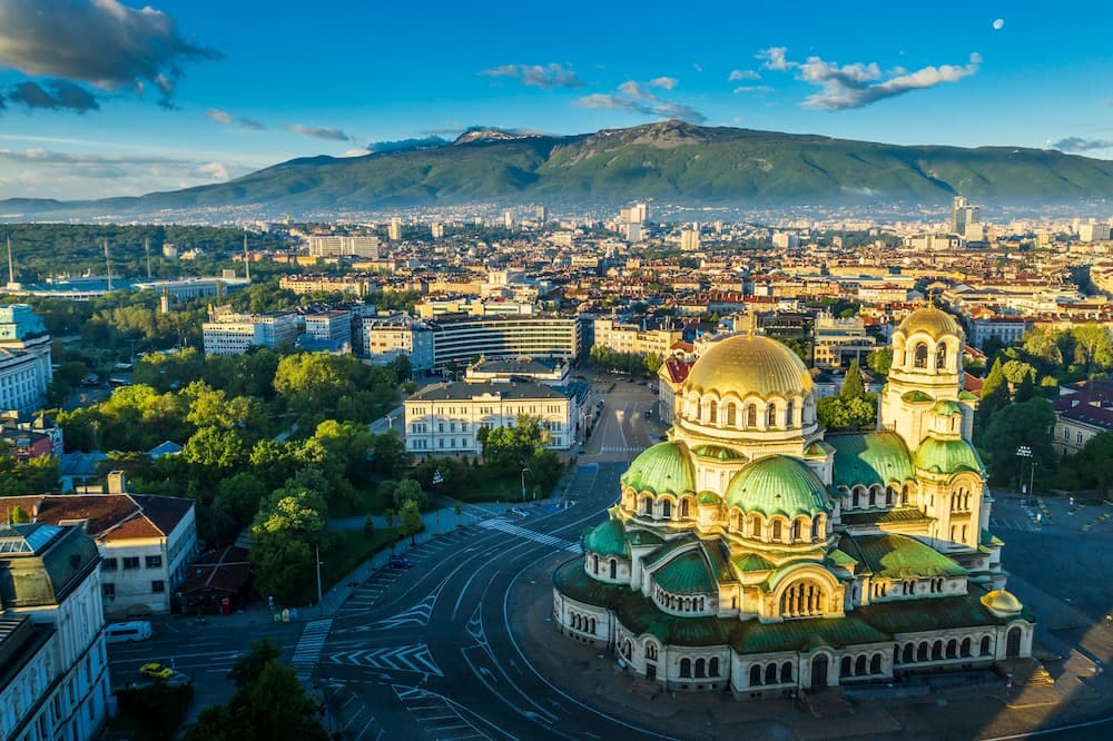 A panoramic aerial view of Sofia, Bulgaria, featuring the Alexander Nevsky Cathedral with its large gold and green domes in the foreground, surrounded by city streets, trees, and historic buildings, with Mount Vitosha rising in the background under a clear blue sky.