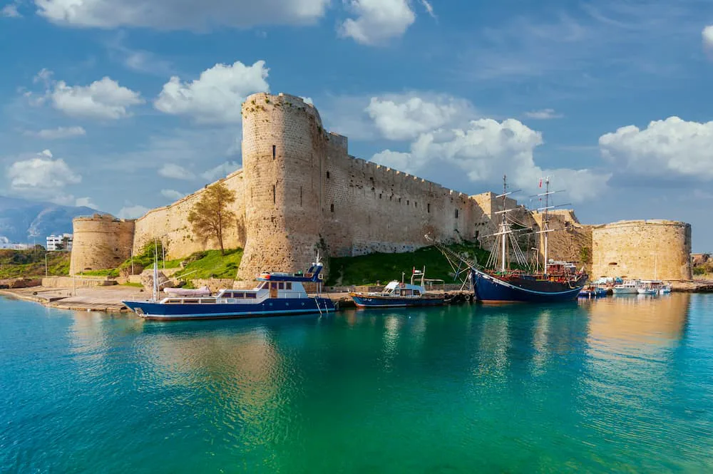 A historic stone fortress on the coast of Cyprus overlooking a calm harbour, with small boats and a wooden sailing ship moored in turquoise water beneath a partly cloudy blue sky.