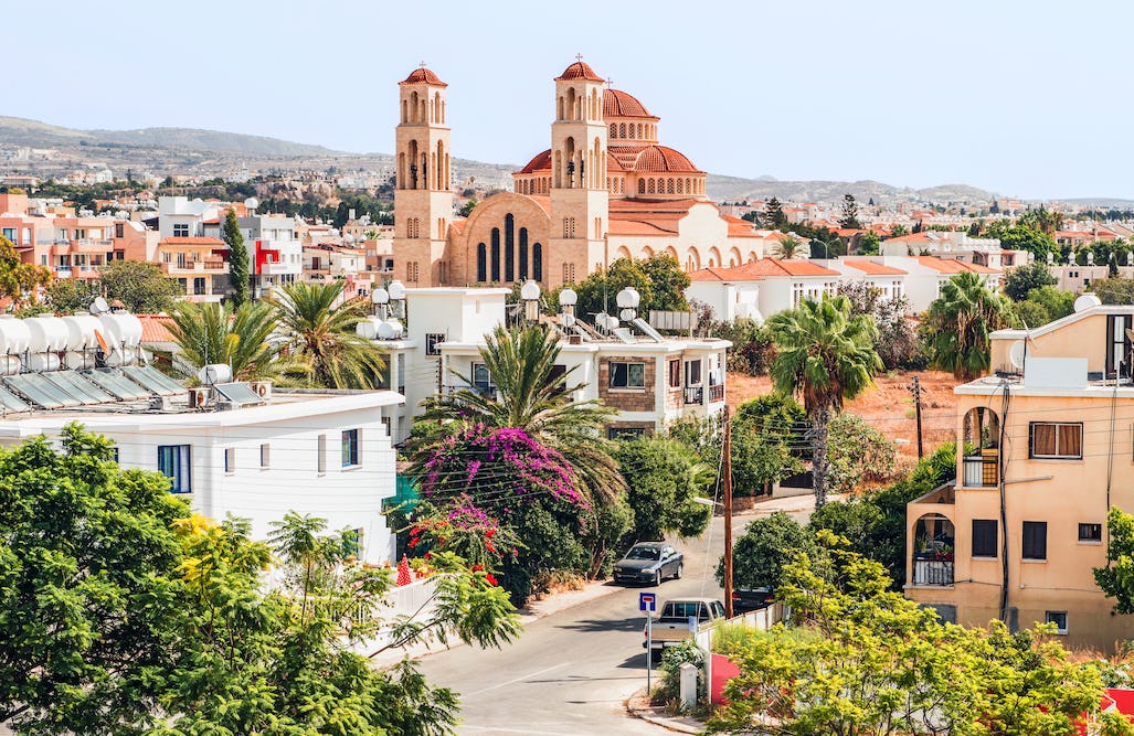 A residential neighbourhood in Cyprus with palm trees and low-rise homes, featuring a large red-tiled church with twin bell towers rising above the buildings, set against rolling hills under a clear sky.