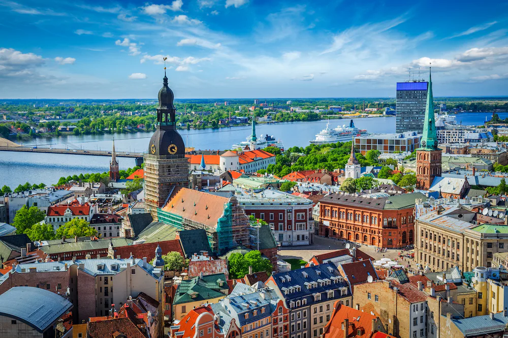 A panoramic view of Riga, Latvia, showing the historic Old Town with colourful rooftops, church towers, and the River Daugava in the background, with boats on the water and green riverbanks under a bright blue sky.