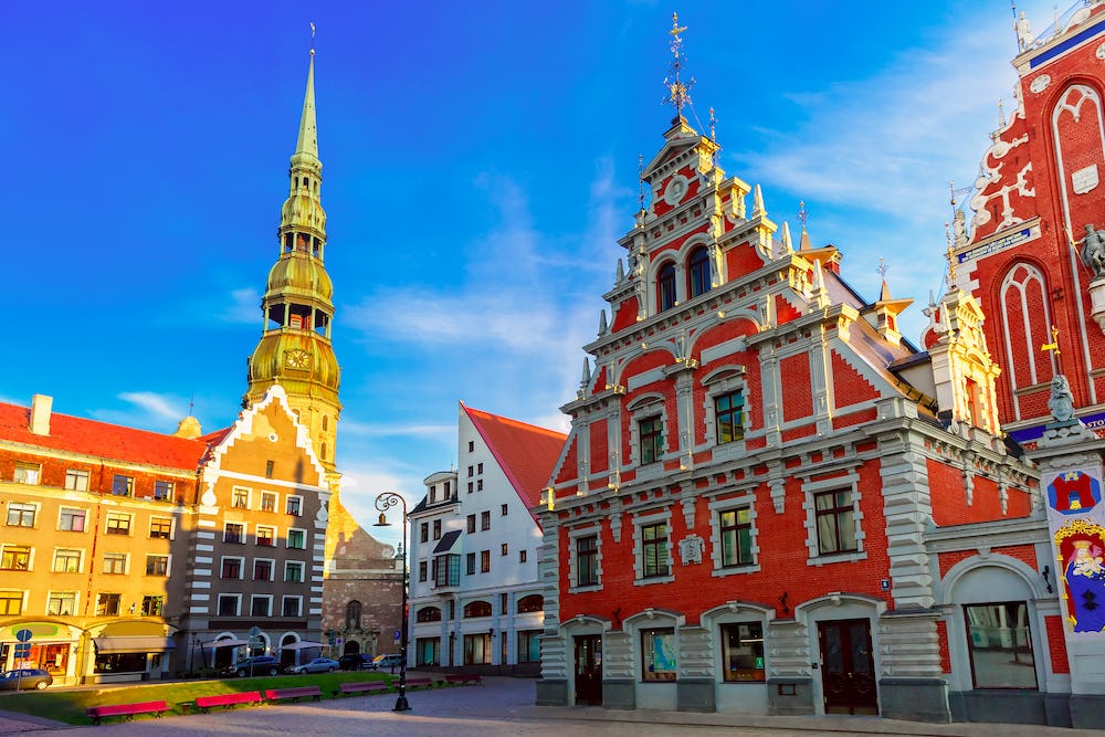 A sunlit view of Riga’s Old Town square in Latvia, featuring ornate red-brick guild buildings and a tall church spire, with cobblestone streets and historic architecture beneath a clear blue sky.