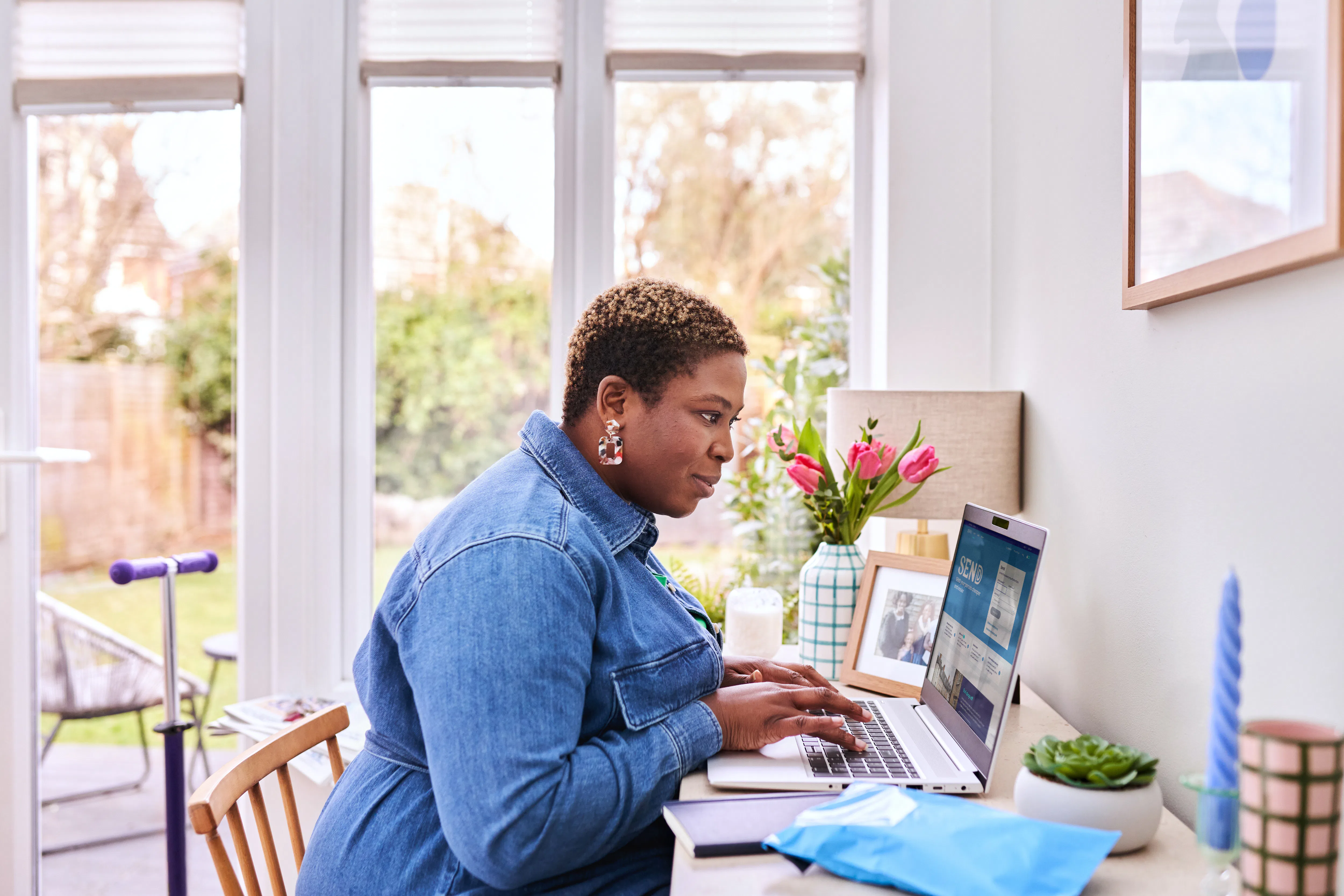 woman sat at counter using laptop with parcel next to her and other household items