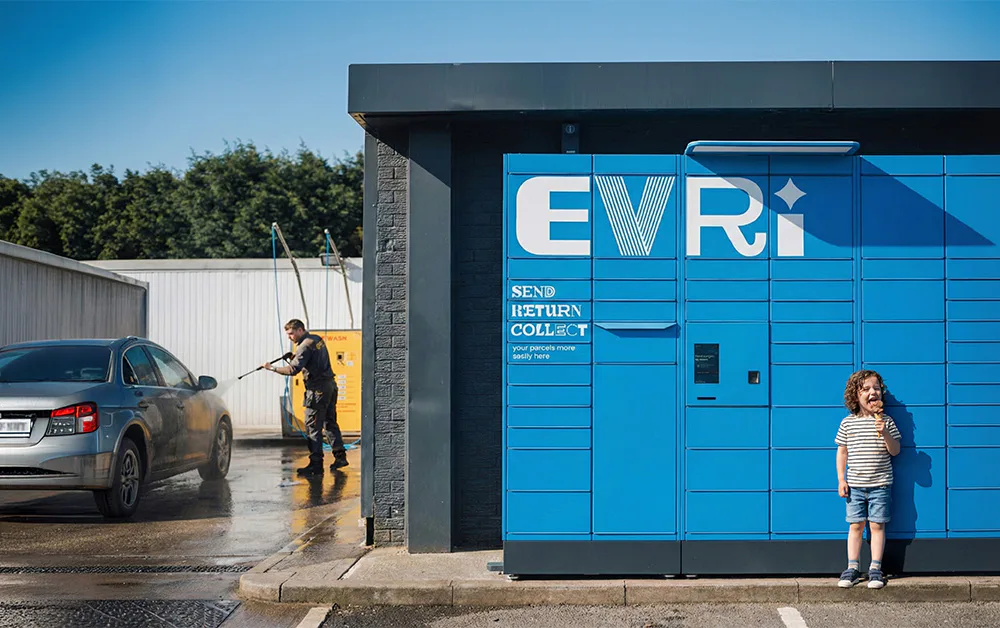 A bright blue Evri parcel locker stands beside a self-service car wash, where a person is washing a silver car. A child holding an ice cream stands smiling next to the locker in the sunlight.