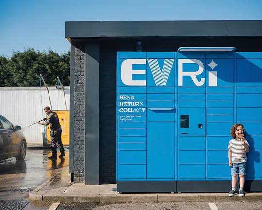 A bright blue EvrRi parcel locker stands beside a self-service car wash, where a person is washing a silver car. A child holding an ice cream stands smiling next to the locker in the sunlight.