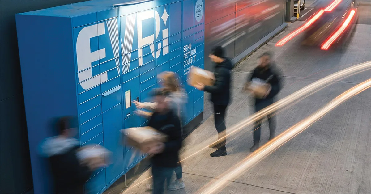 Multiple people collect and return parcels at a blue Evri Locker while traffic light trails pass by on a nearby road at dusk.