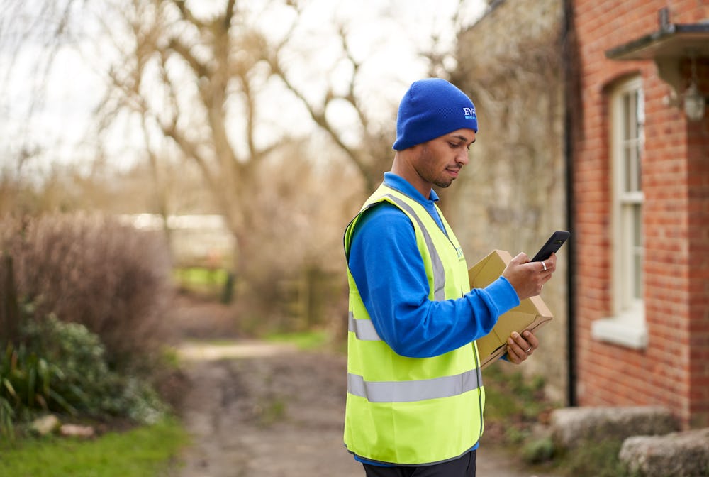Photo of a courier in hat holding package and smartphone outide house