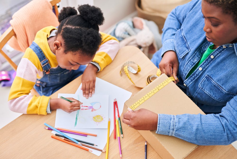 woman holding tape measure to cardboard box while child draws sitting next to her