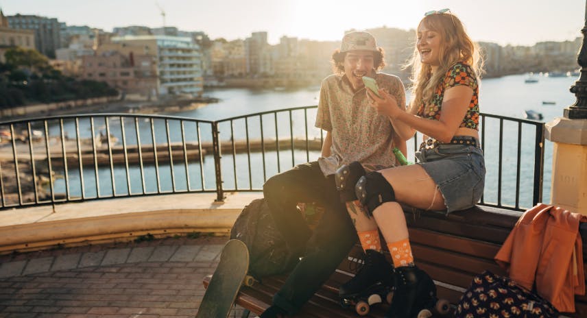 Skater sitzen auf einer Parkbank in ihrer Stadt und lachen, während sie sich Fotos anssehen, die sie gerade mit einem Smartphone aufgenommen haben