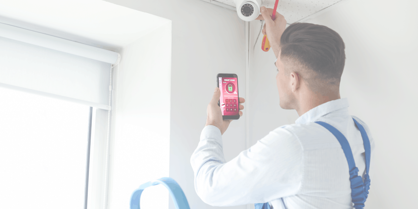 Young male installer dressed in a white shirt and a blue overall installing a smart surveillance camera and holding a smartphone to remotely control the security system