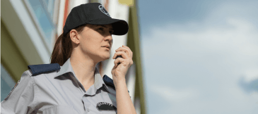 Female security guard with a ponytail, uniform, and black base cap speaks into a walkie-talkie outside a company building during daytime