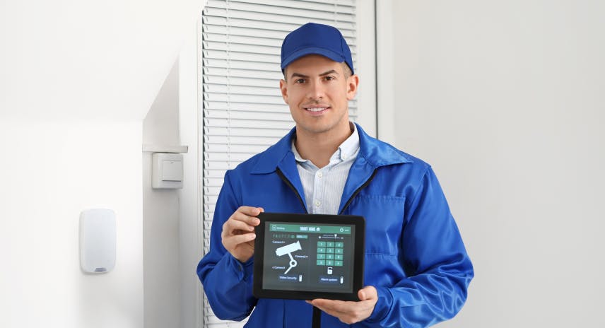 Männlicher Sicherheitstechniker in blauer Kleidung mit Basecap, der lächelnd ein Tablet hält, auf dem eine intelligente Überwachungskamera abgebildet ist