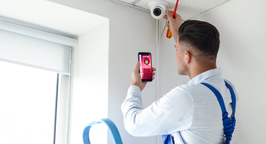 Young male installer in a white shirt and blue overall installing a surveillance camera and holding a smart phone to remotely control the security system