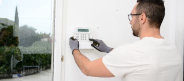 Security technician wearing a white t-shirt, gloves, and glasses is installing an alarm panel on a white wall next to a big window