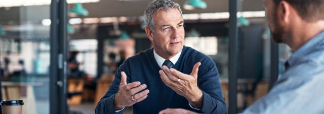 Well-dressed middle-aged businessman in an open office space gesticulating with open hands and talking to a business partner