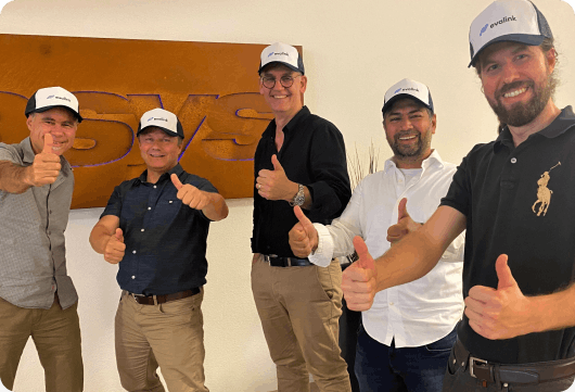 Foto eines glücklichen evalink-Teams in der Sitasys-Zentrale, alle zeigen Daumen nach oben, lächeln und tragen blau-weiße evalink-Basecaps