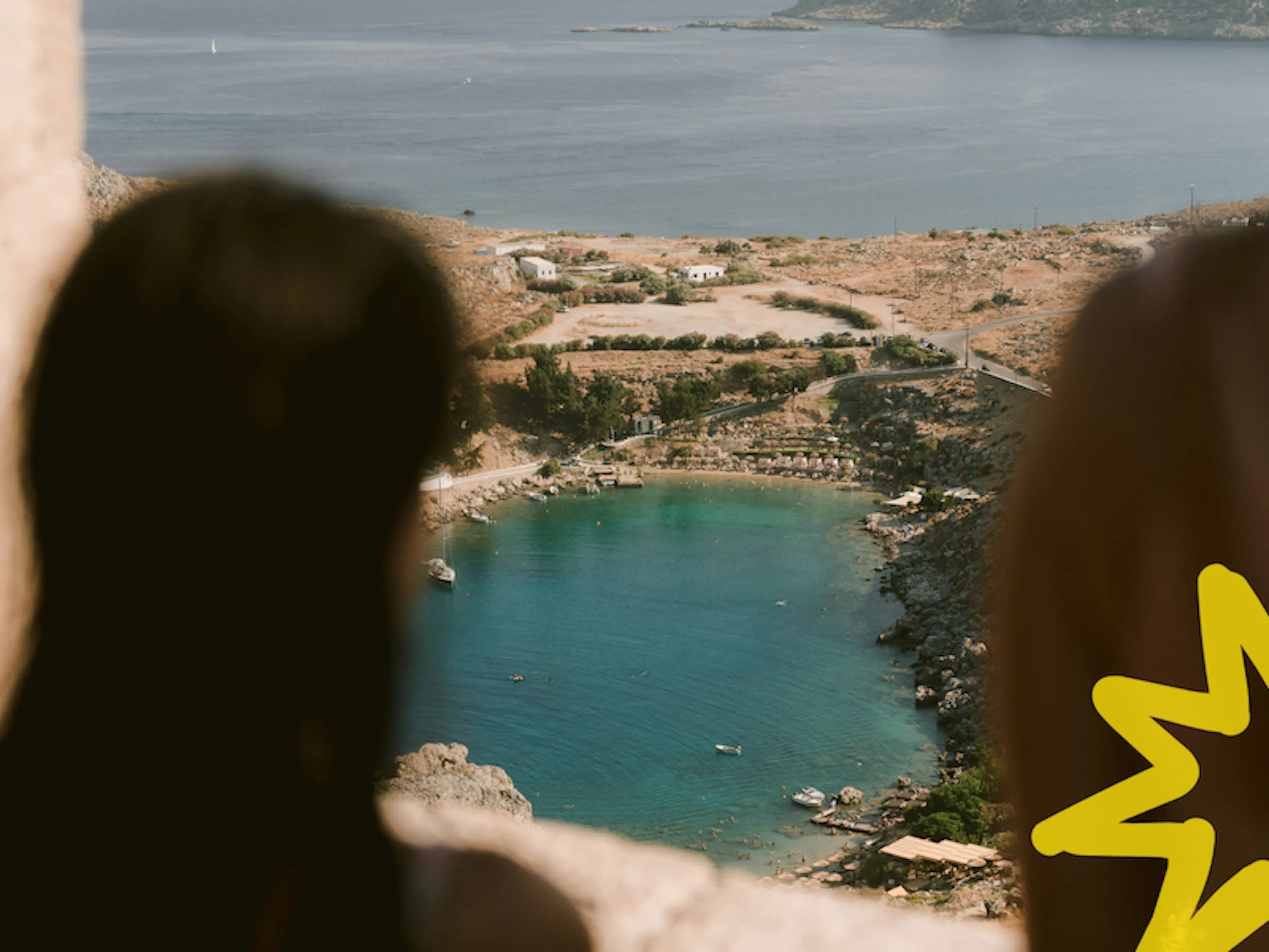 Dos mujeres contemplando desde lo alto una playa de aguas turquesas en Grecia