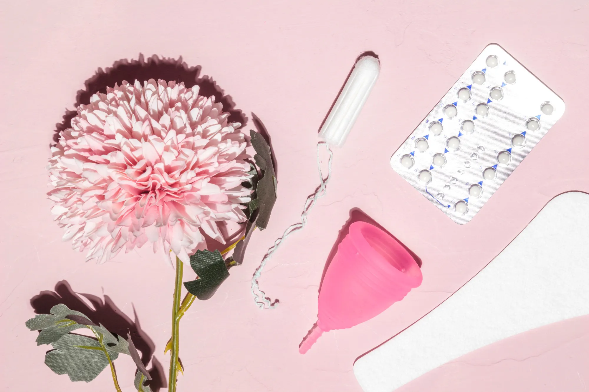 Flat lay featuring a flower, menstrual cup, tampon, and birth control pills, representing the female cycle and choices in reproductive health and contraception.