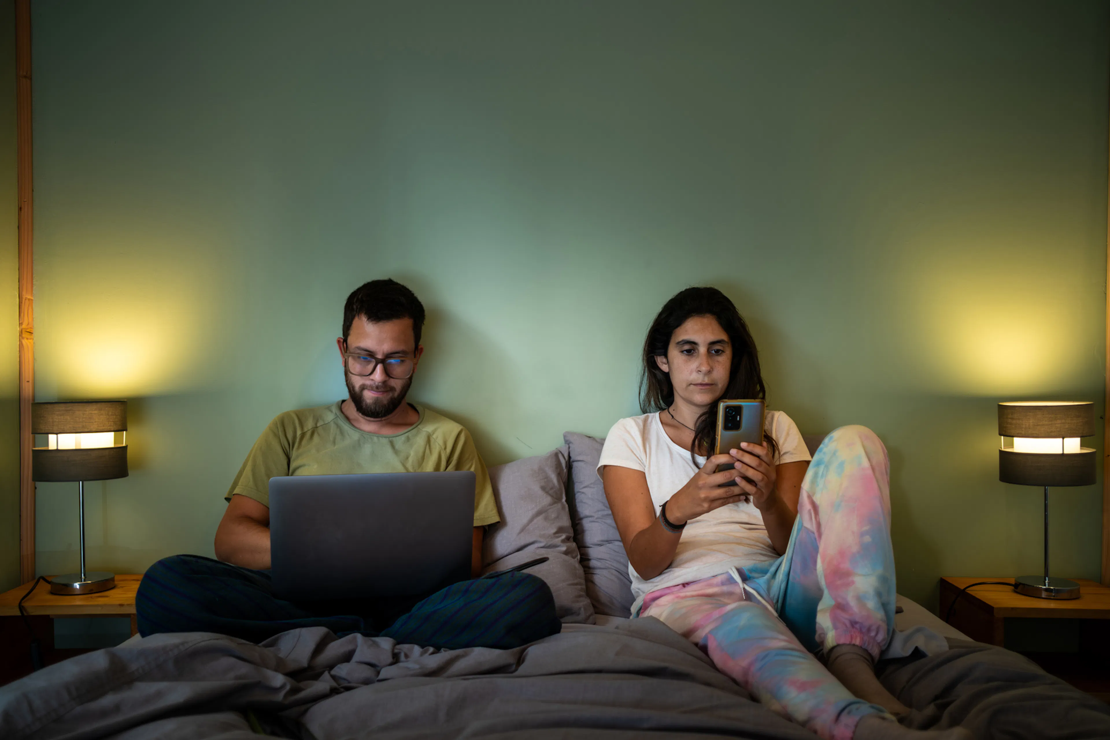A woman and a man are sitting in bed. The woman is looking through her phone, while the man is on his laptop. There's space between them. 
