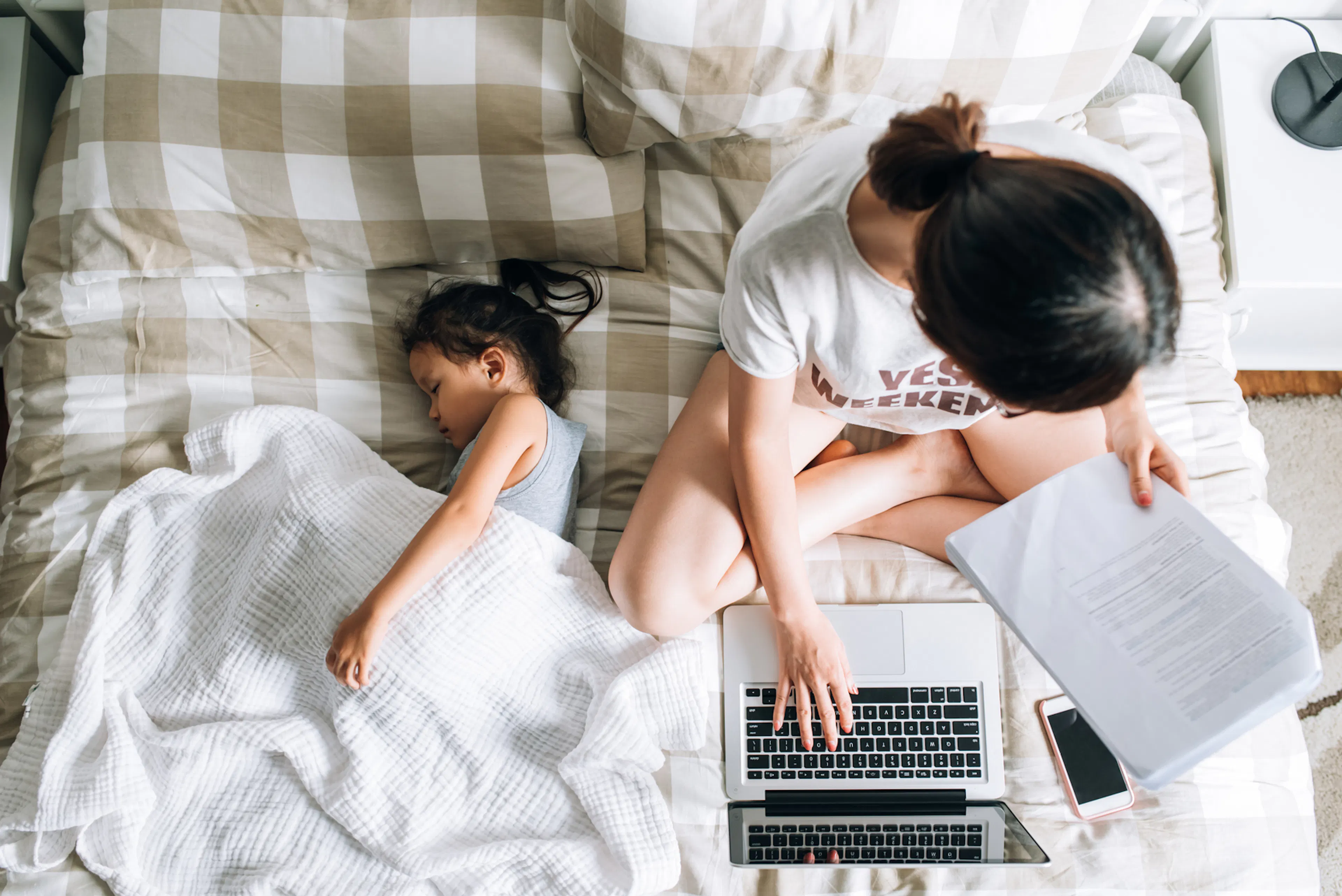 Mom and child are in bed. Mom is looking at paperwork while child sleeps.