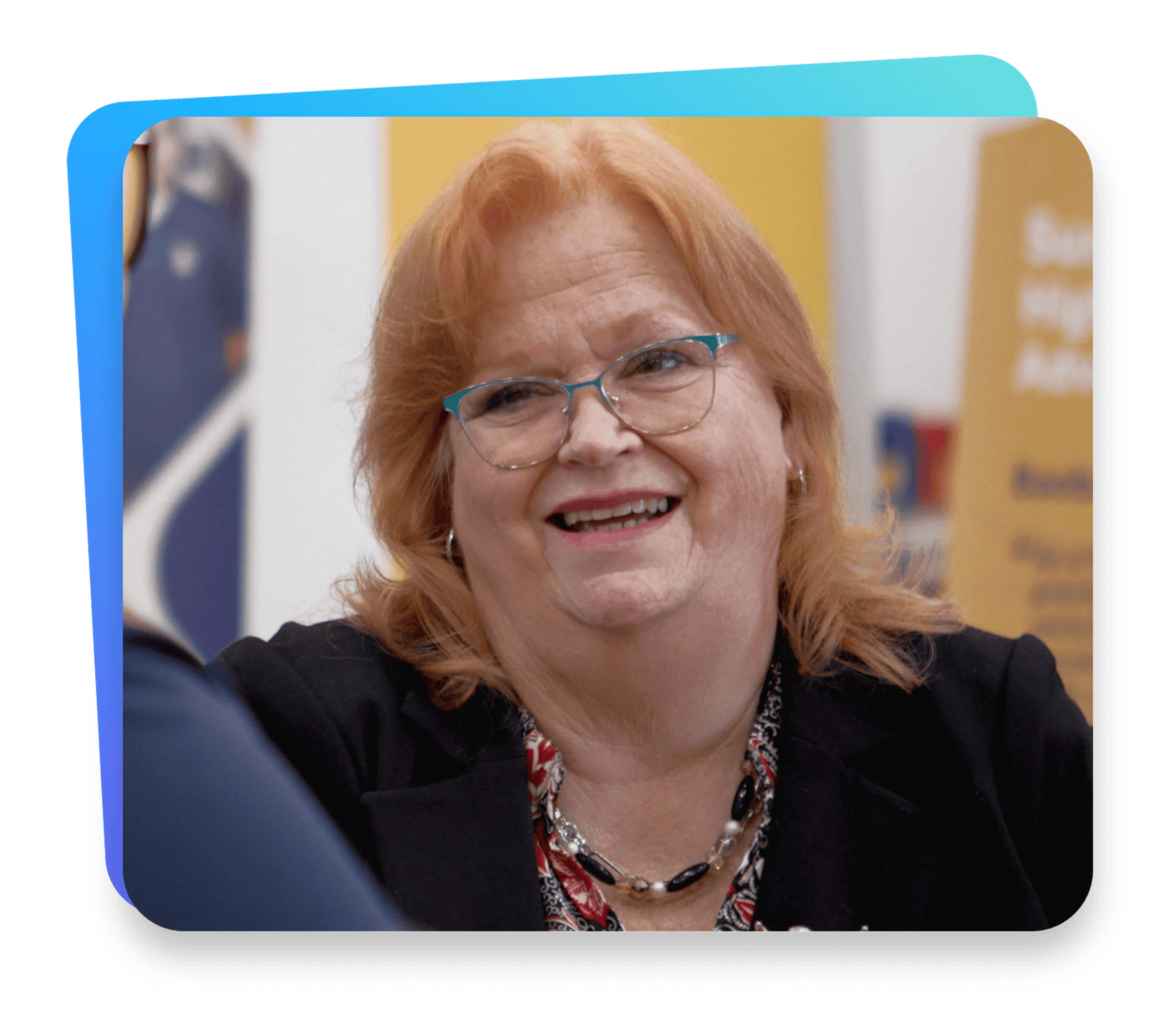 Smiling woman with red hair and glasses wearing a black blazer, engaged in conversation at a professional event.