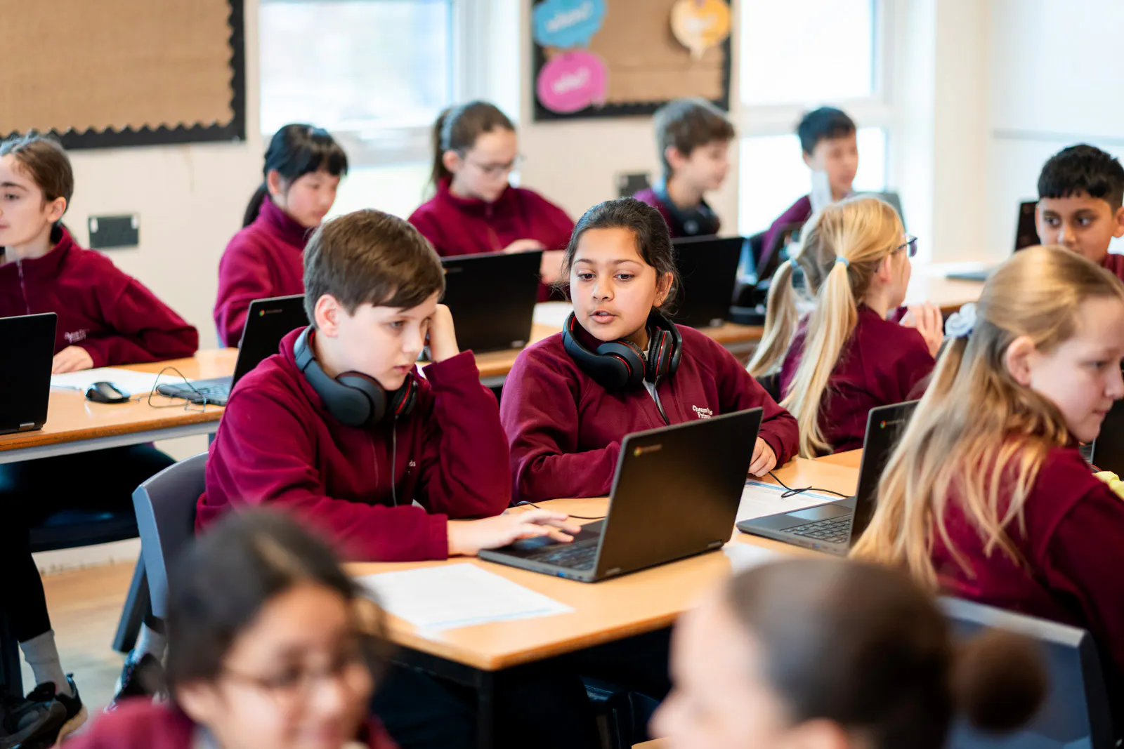 Students collaborate and work independently on laptops in a classroom, with several wearing headphones during a technology-based lesson.