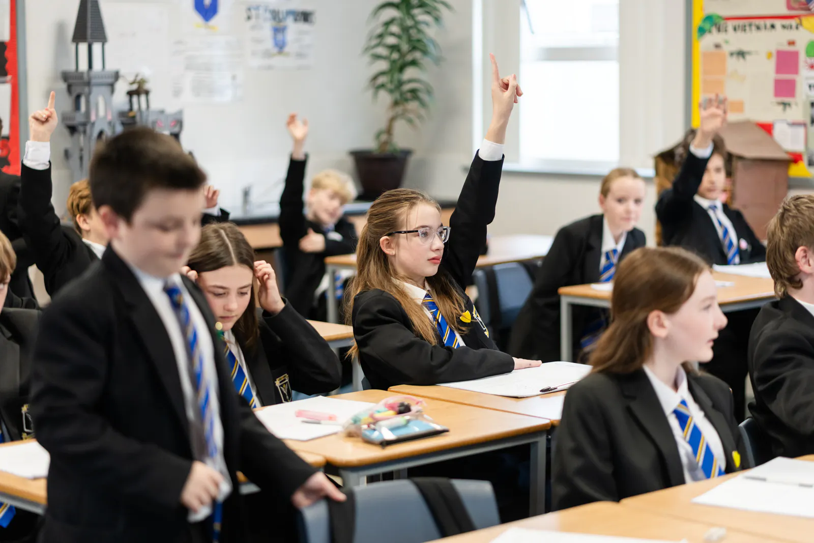 Students raise their hands to answer questions during a classroom lesson, showing active participation and teacher-led discussion.