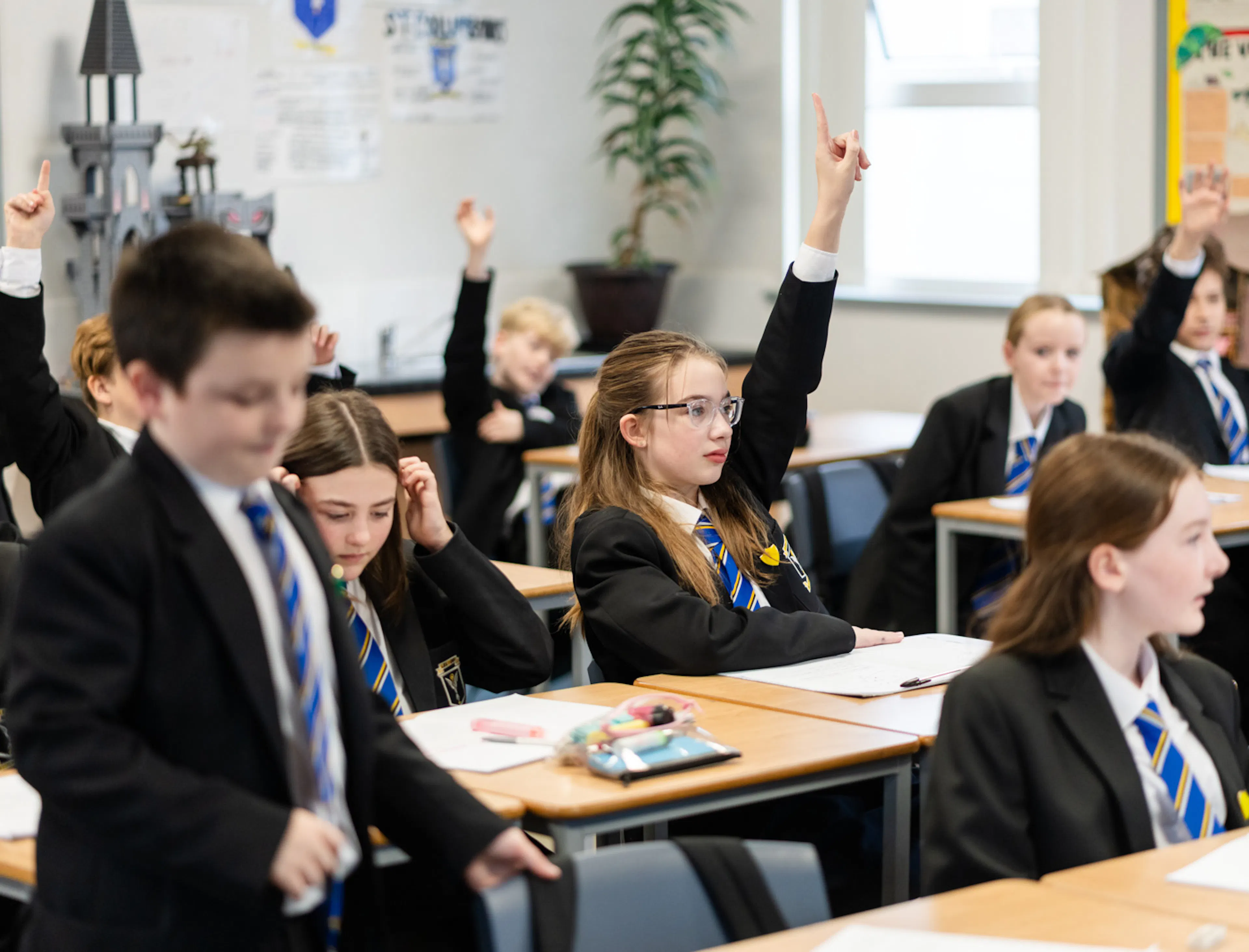 Students raise their hands to answer questions during a classroom lesson, showing active participation and teacher-led discussion.