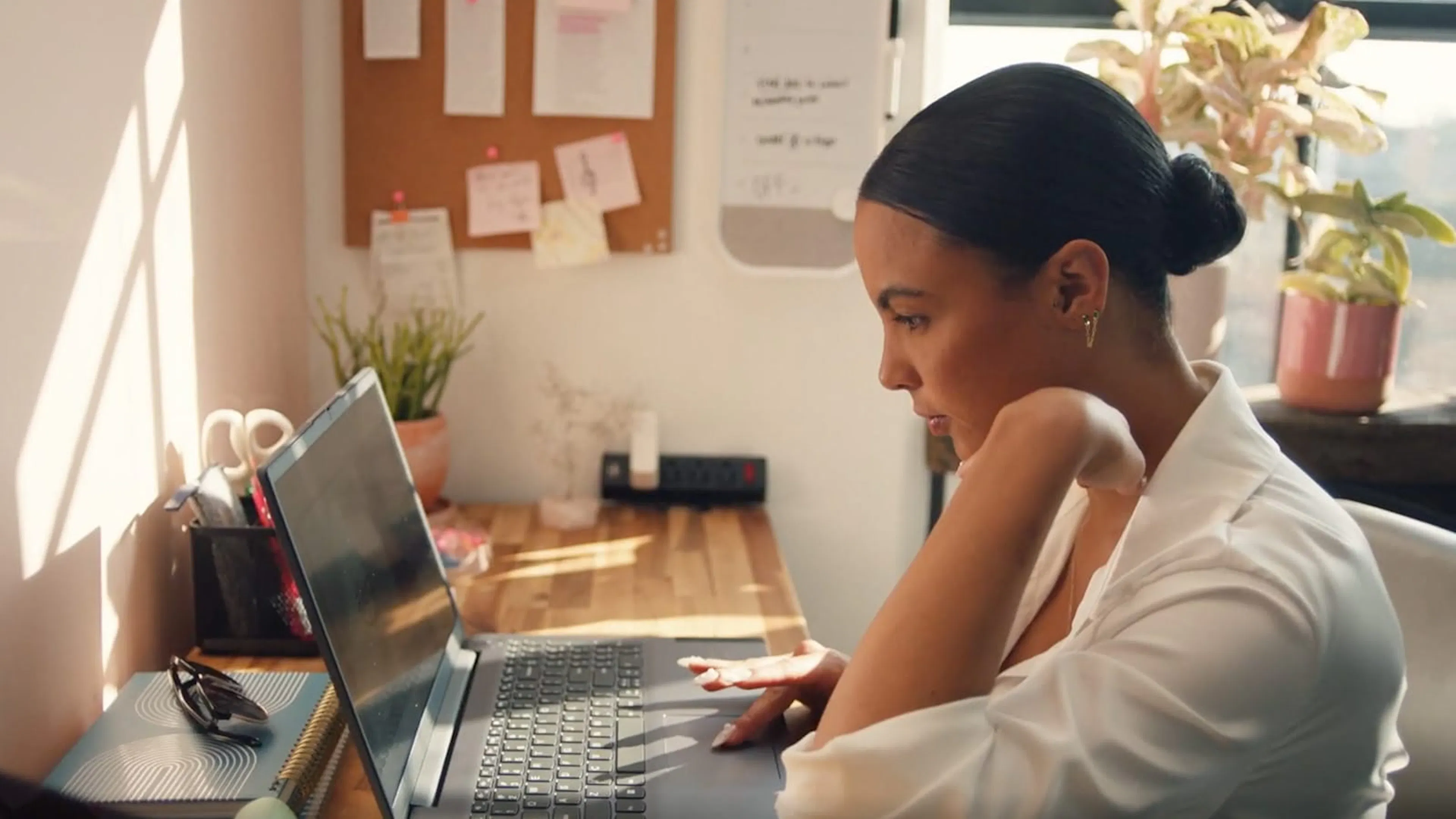 A woman (Eva) in a white shirt sits working on a laptop at a sunlit desk, surrounded by plants and office notes.