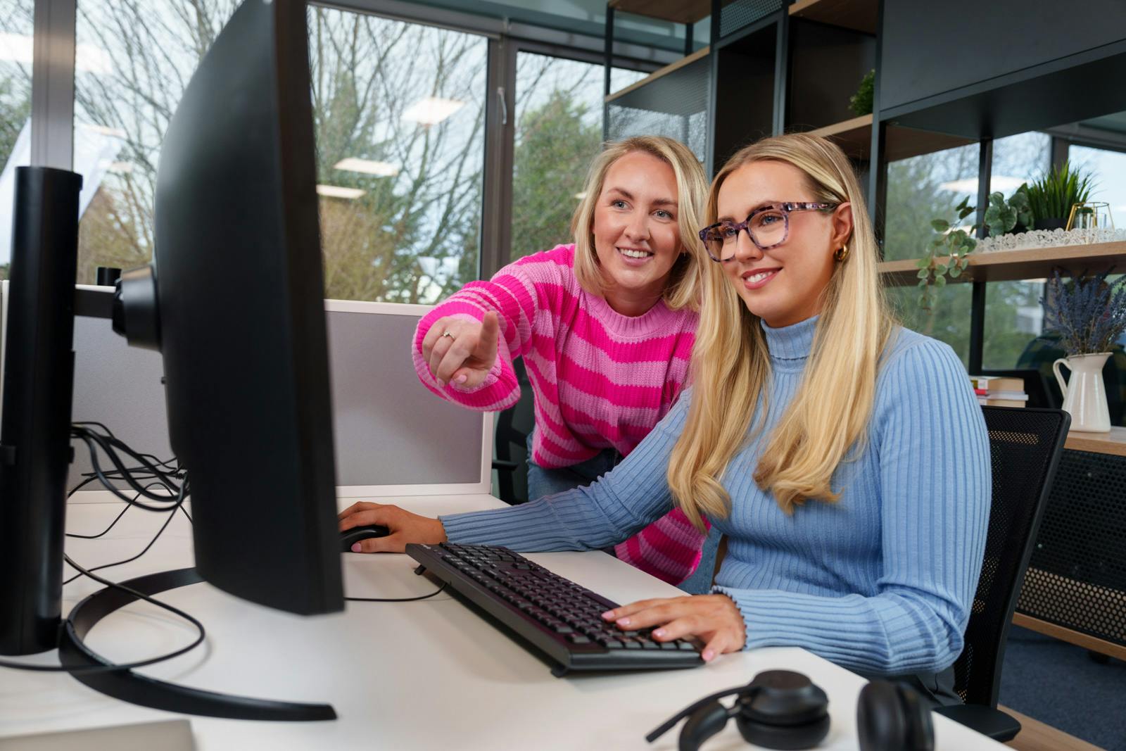 Two office employees are working together whilst looking at a screen, one is pointing.