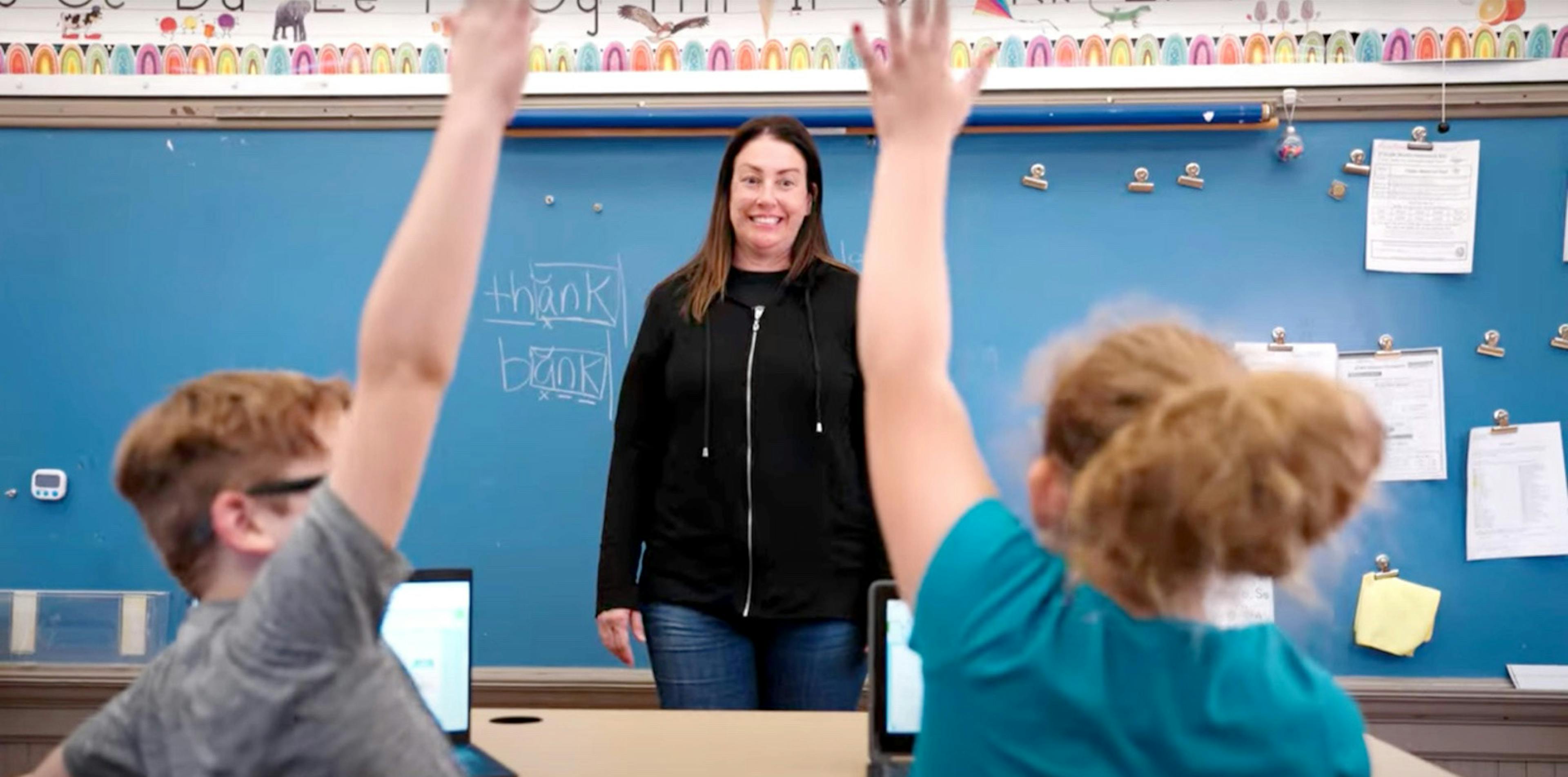 A teacher standing infront of the class while two student raise their hand