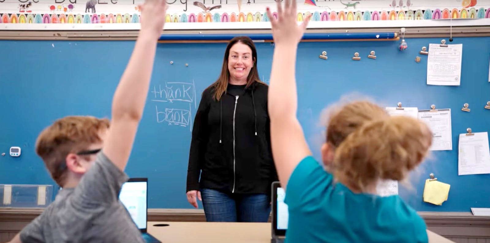 A teacher standing infront of the class while two student raise their hand