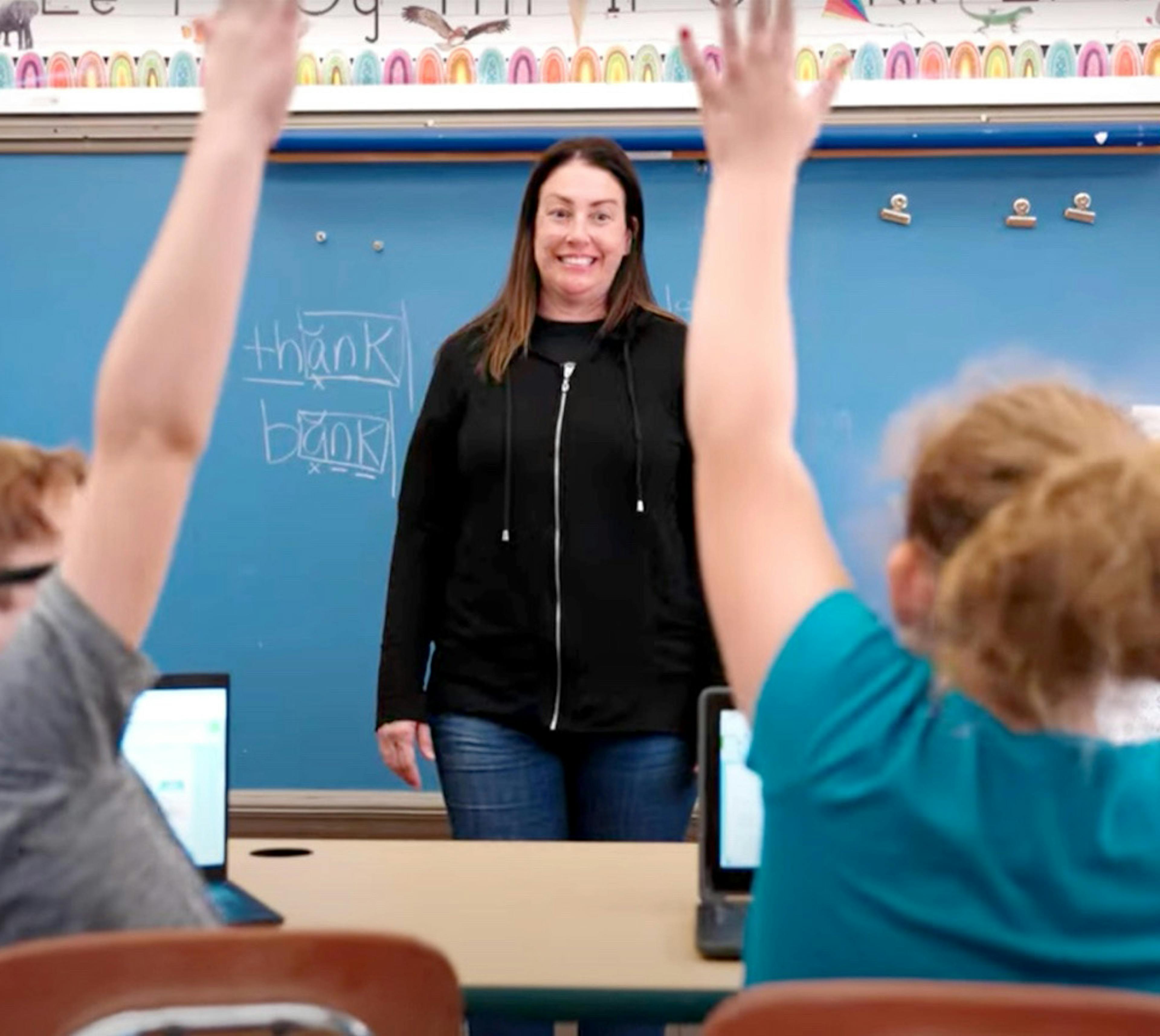 A teacher standing infront of the class while two student raise their hand