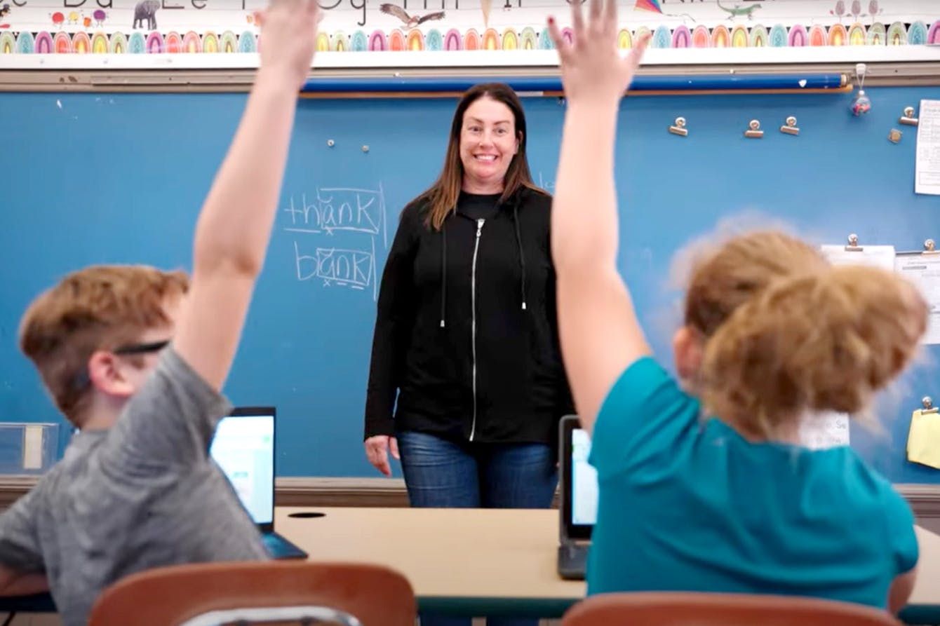 A teacher standing infront of the class while two student raise their hand