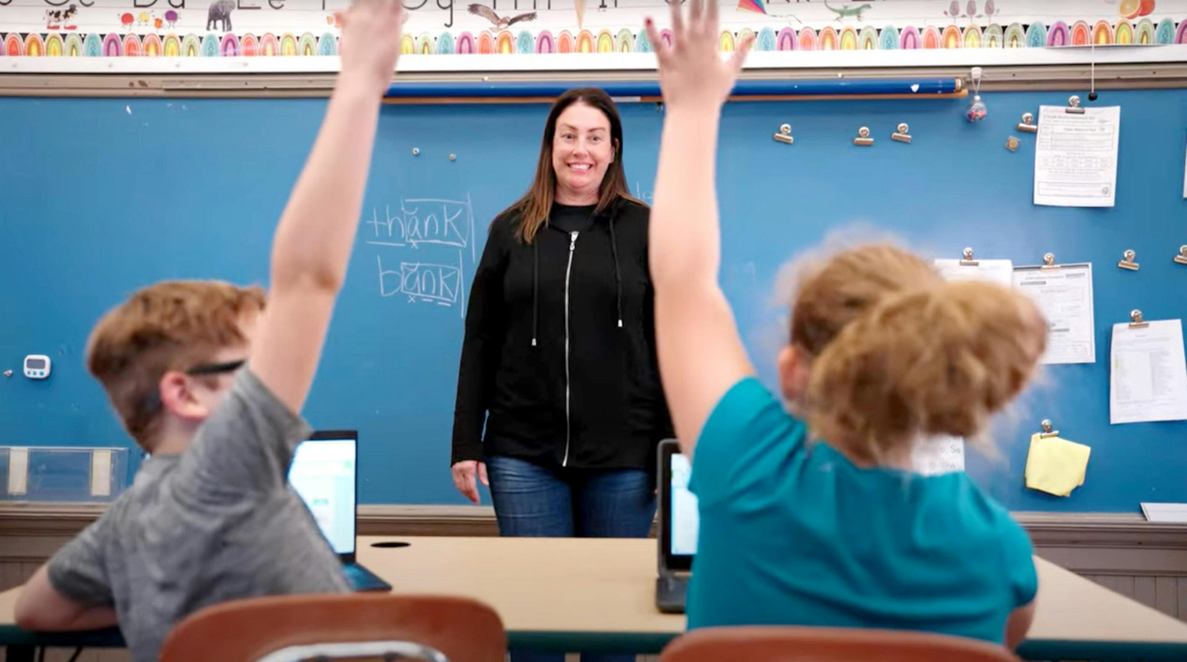 A teacher standing infront of the class while two student raise their hand