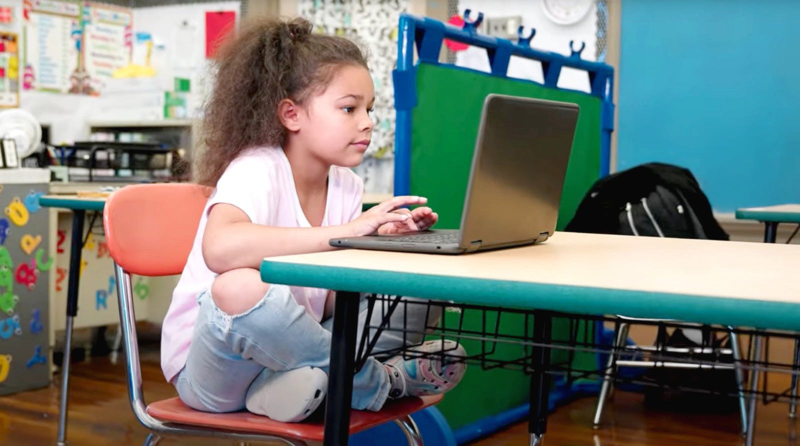 Young student sitting crossed legged on her chair in a classroom. She is typing on a laptop.
