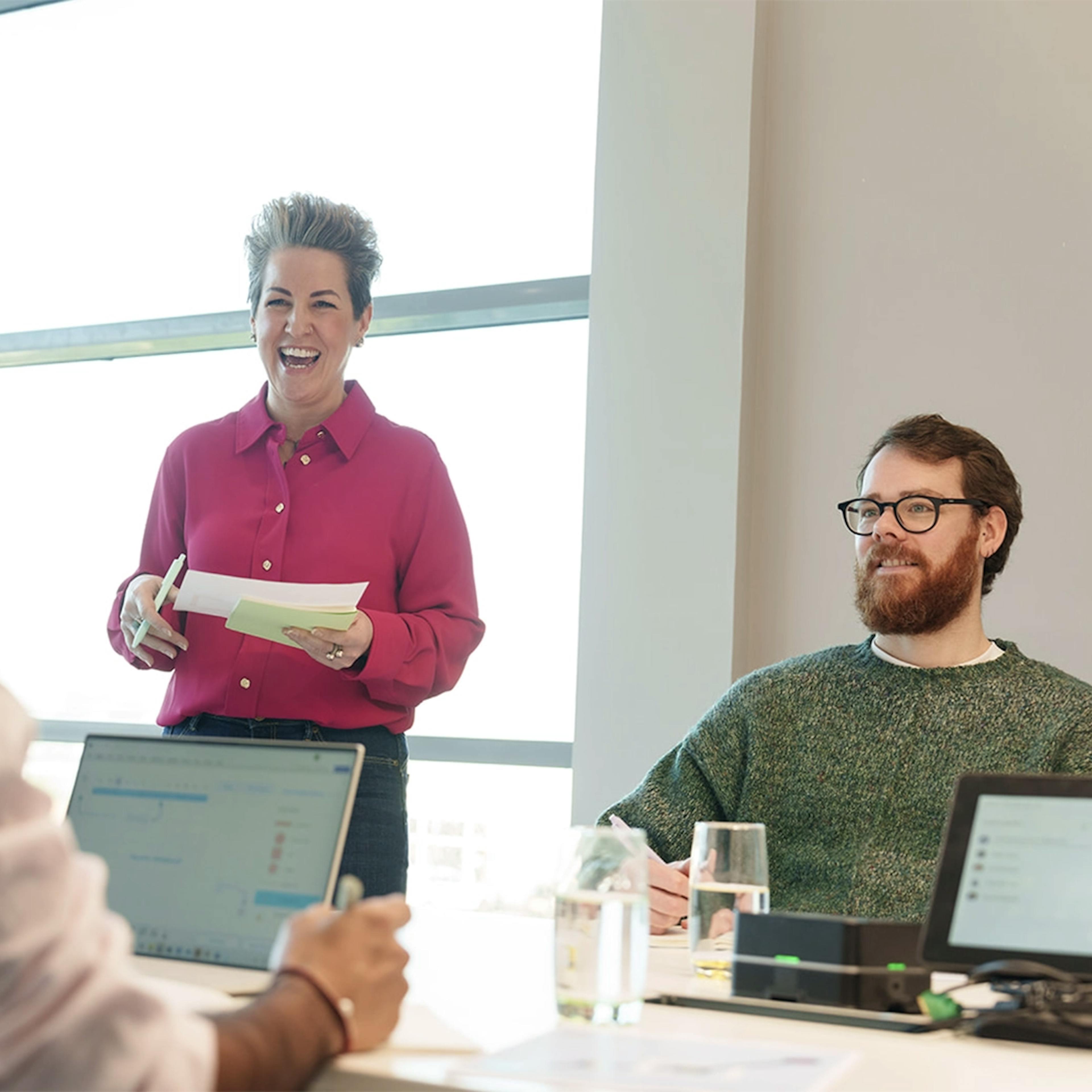 Three employees in a meeting room chatting with laptops and print outs