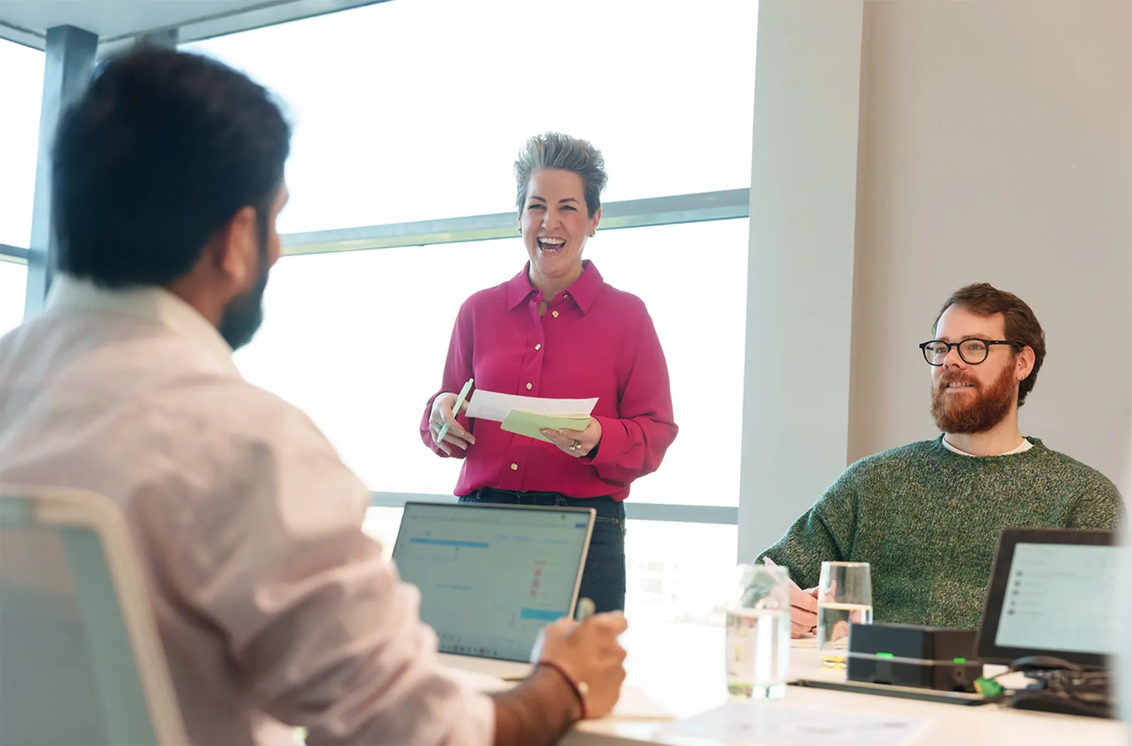 Three employees in a meeting room chatting with laptops and print outs