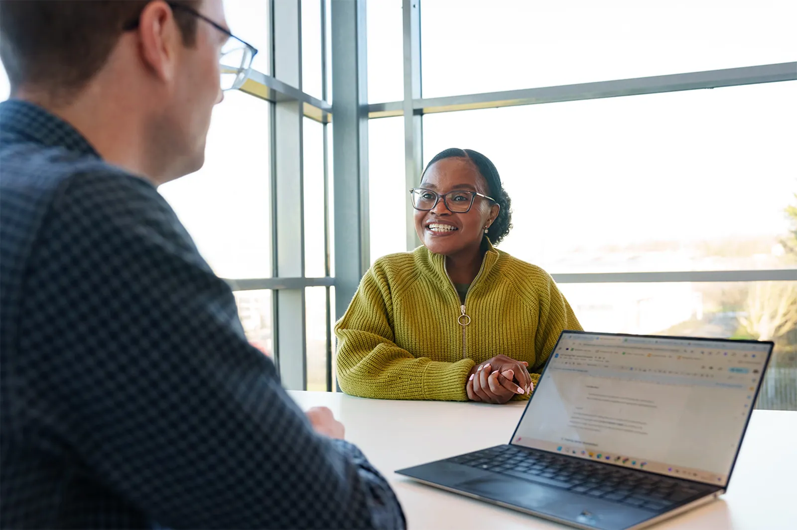 Two professionals sit across a table with a laptop, engaged in a conversation during a meeting in a bright office.