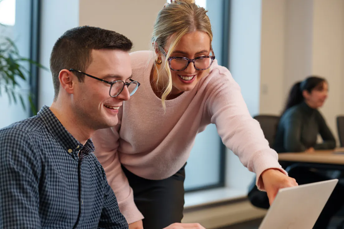 Two smiling colleagues in an office. One sitting at a computer, while the second stands beside them pointing at the computer screen.