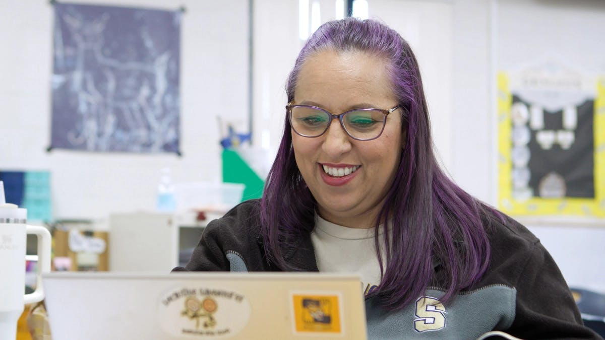 Teacher with purple hair and glasses smiles while working on a laptop in a classroom.