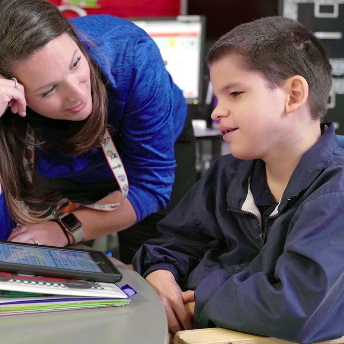 Teacher interacting with a student using a tablet and binder in a classroom setting.