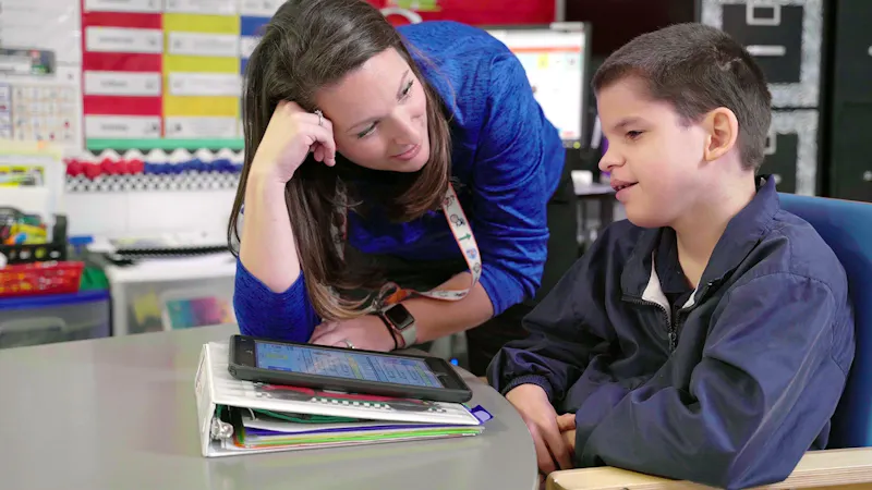 Teacher interacting with a student using a tablet and binder in a classroom setting.