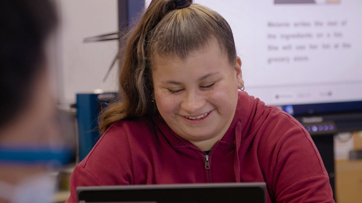 Student smiling while working on a laptop in a classroom.