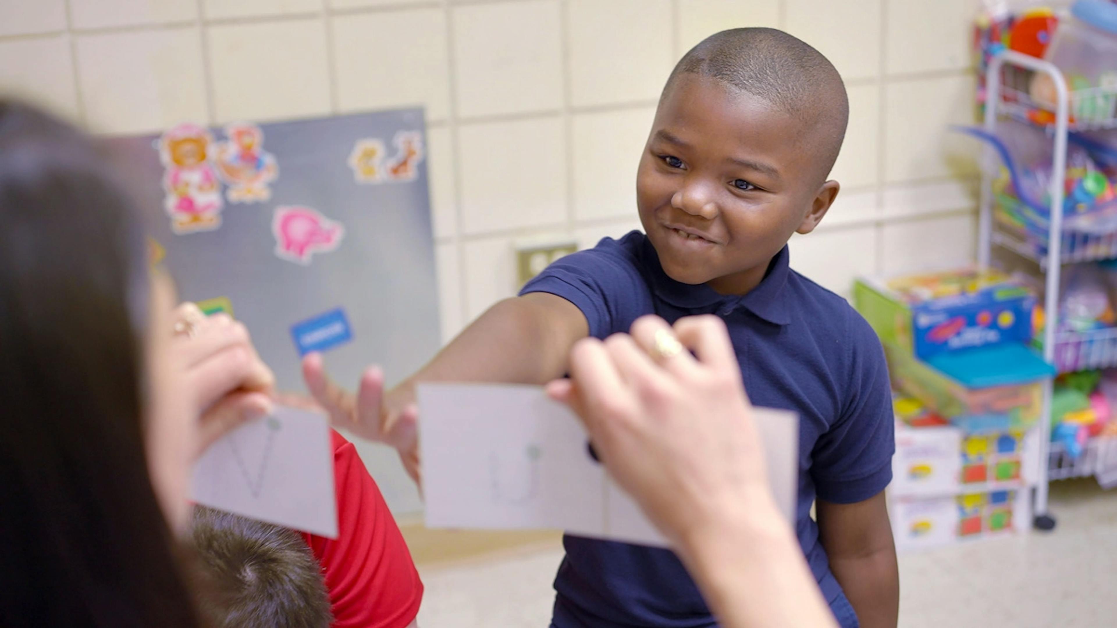 Child smiling while pointing to a flashcard during a classroom learning activity.