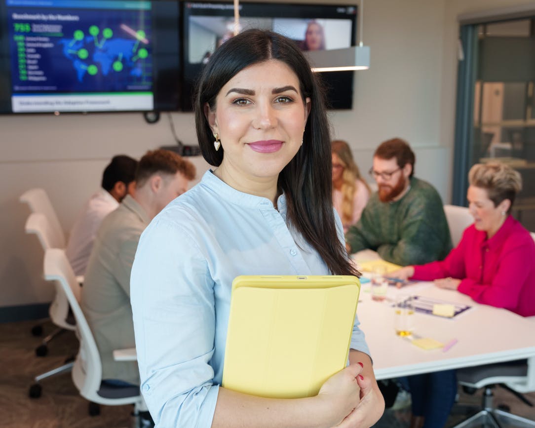 Employee standing in front of a meeting room desk, with others collaborating in the background