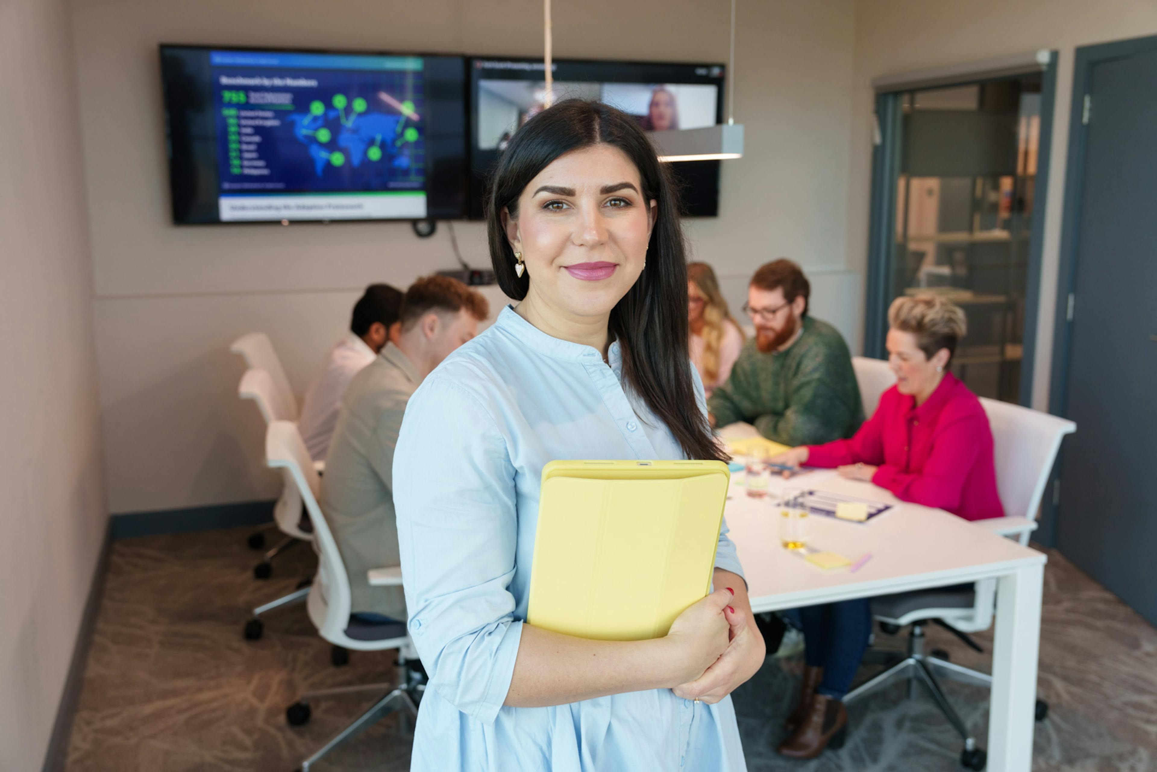 Employee standing in front of a meeting room desk, with others collaborating in the background