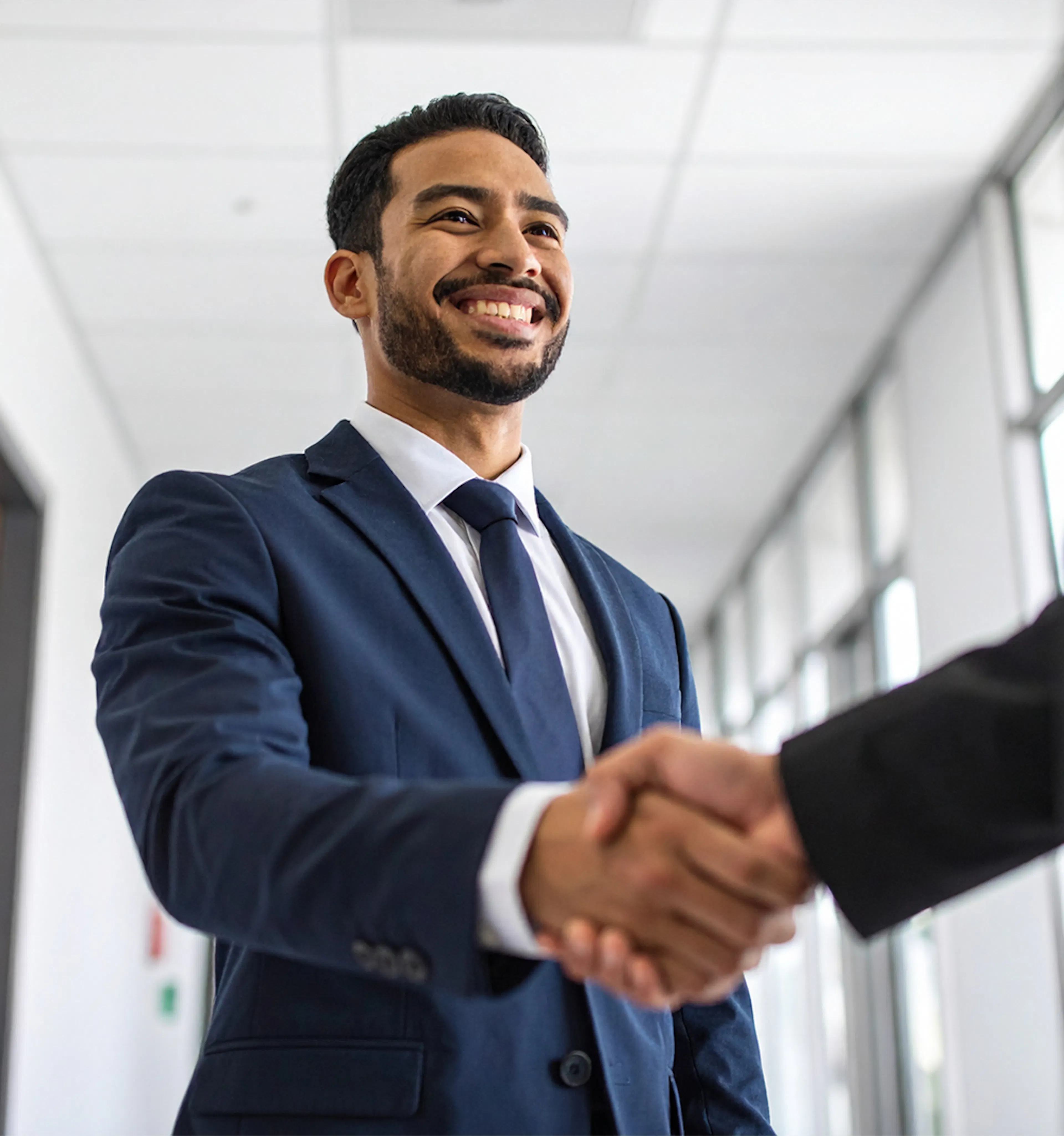 Photograph of a professional man wearing a suit shaking hands with another professional, in a brightly lit hallway.