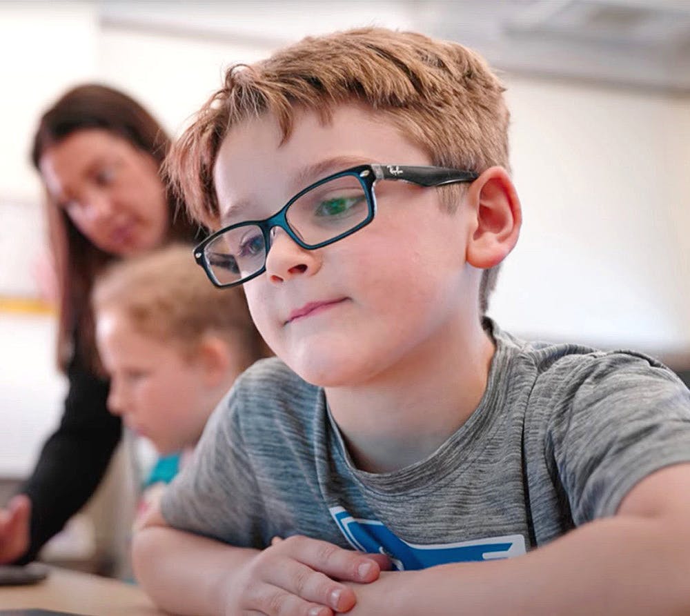 Student working on laptop in classroom. In the background the teacher can be seen assisting another student.