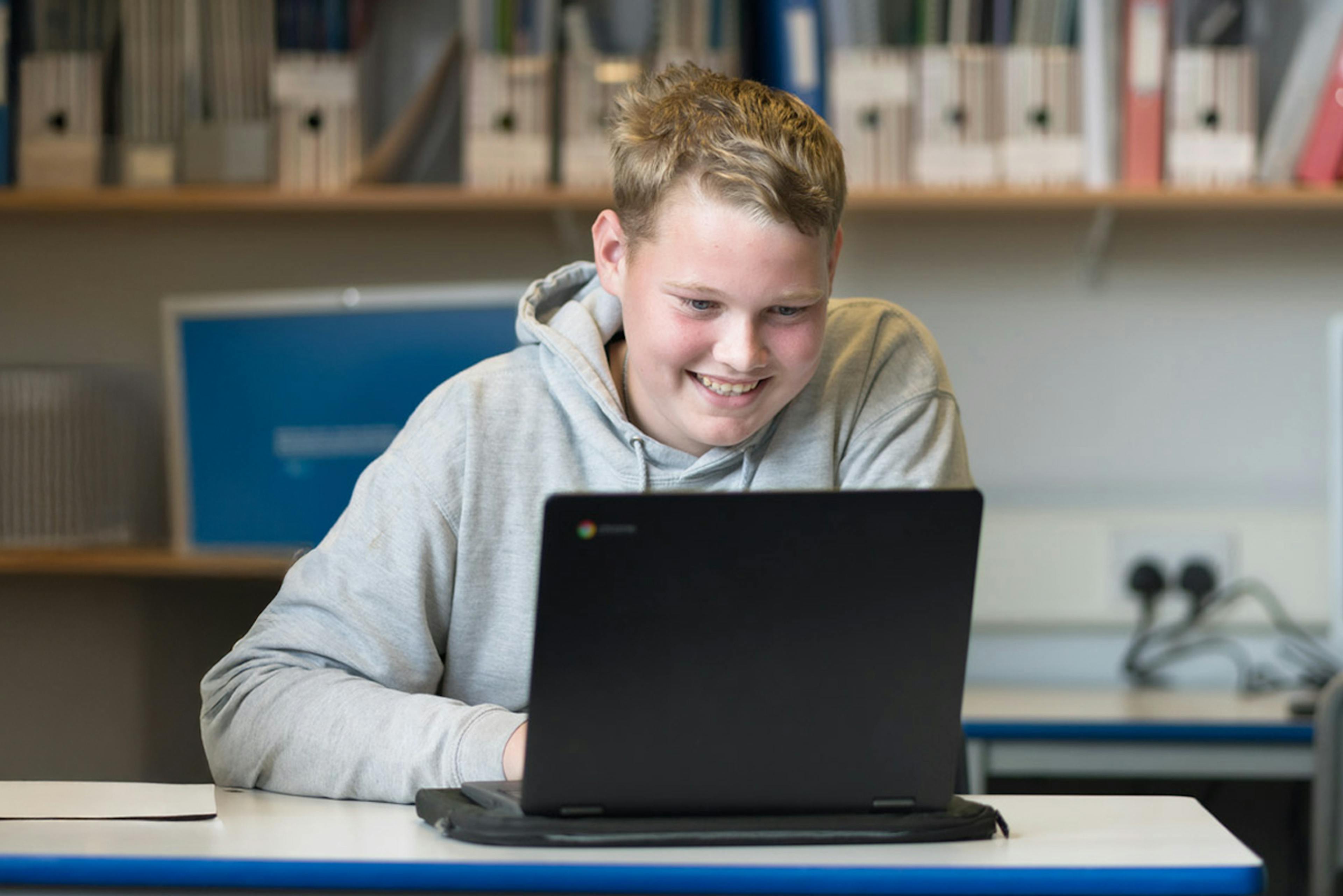 Student in classroom smiling whilst working on a laptop.